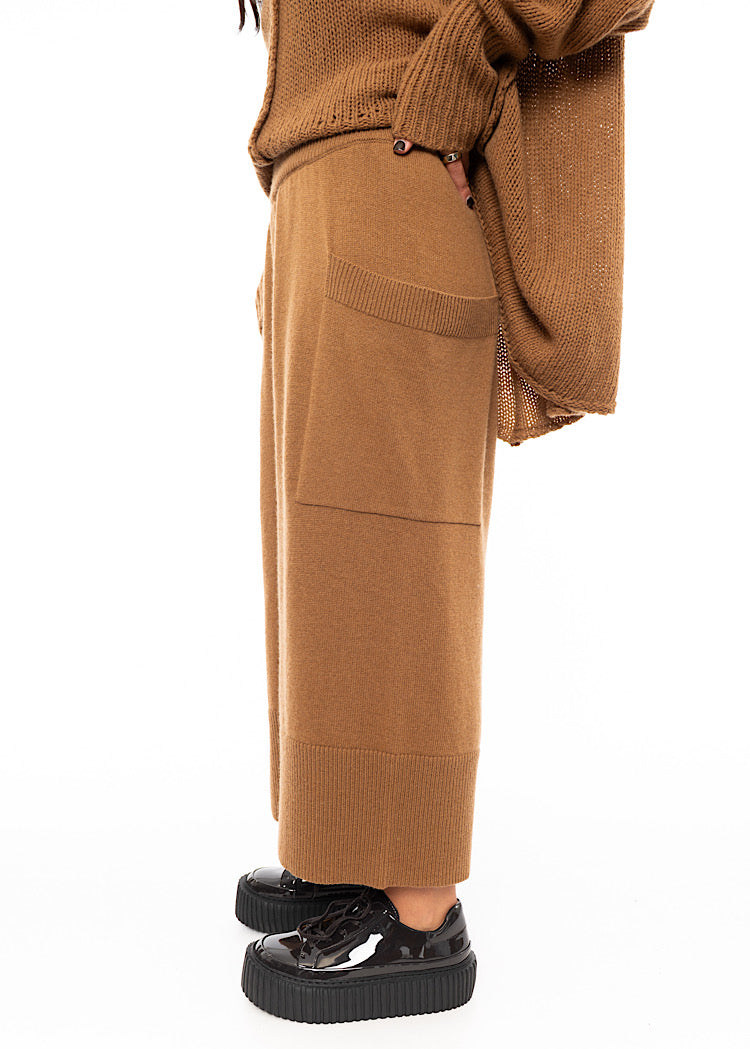 A Model standing to a side wearing a Knitted pair of off-orange Trousers, paired with a matching pullover and patent shoes.