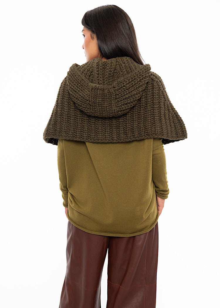A Model facing towards the back wearing an oversized knitted shawl in dark green. Showing how the hood sits on the back.