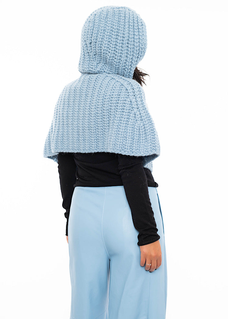 A model standing facing the back wearing an oversized knitted shawl in light blue with hood up. Showing the back length and hood in more detail.