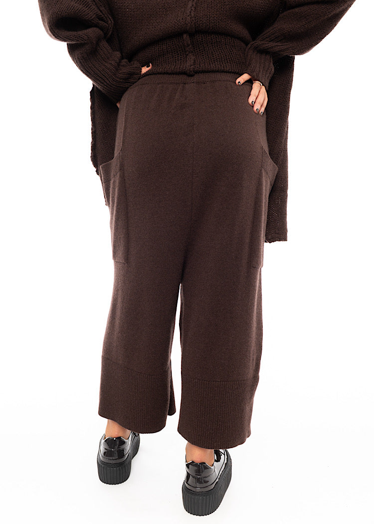 A Model standing facing the back wearing a Brown Knitted Skirt with pockets. Paired with a matching coloured Pullover and Brown patent shoes.