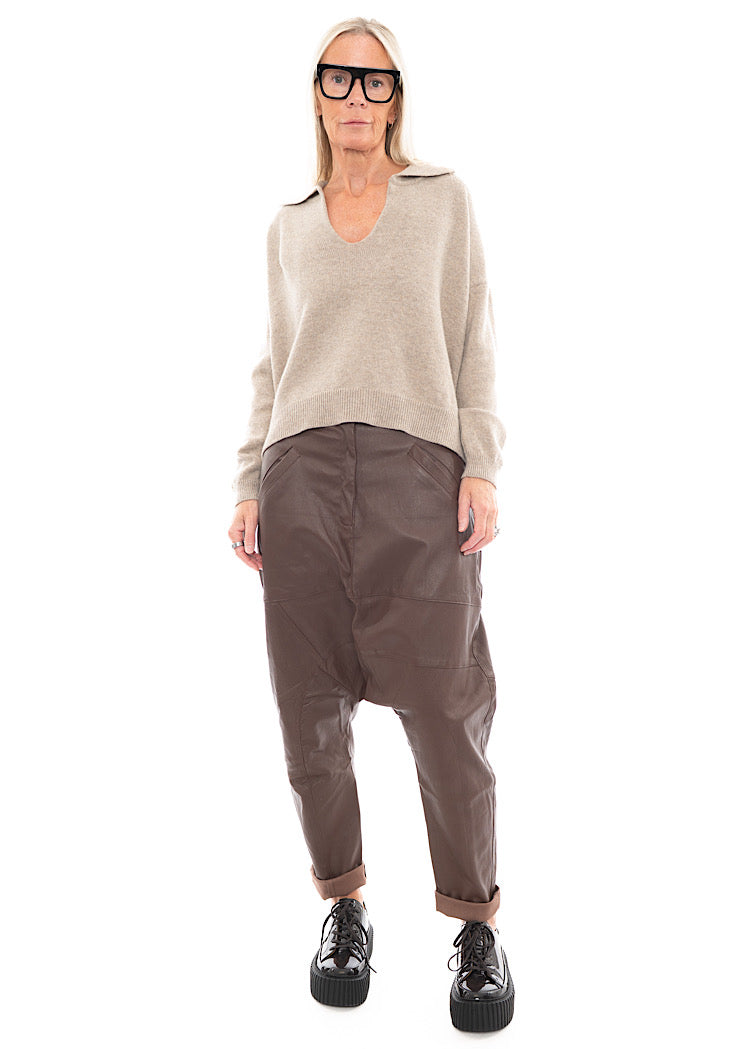 A full frame photo of A Model wearing a Structured Drop-Crotch Brown Leather Trouser.Styled With a patent Brown Shoe and a creme pullover.