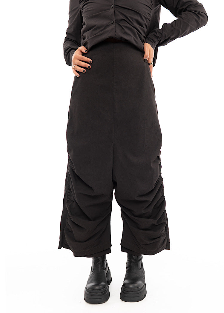 A Model wearing a drop-crotch Trouser in a dark Brown colour. With a kick-flare leg, paired with a matching straight fit shirt.