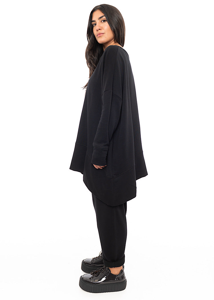 A Full frame photo of A Model standing to a side wearing a Black Oversized Pullover. Showing the oversized but straight fit of the piece.