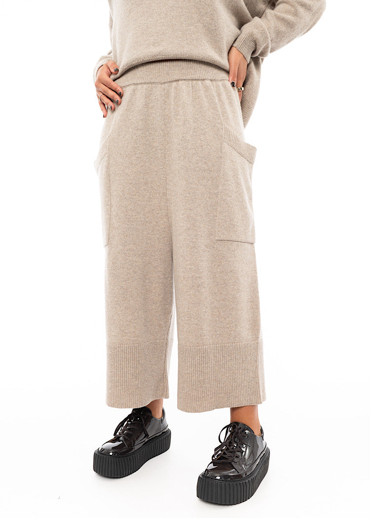 A Model wearing a light beige knitted Trouser. Paired with a matching pullover and Brown patent Shoes.Showing the waistband of the piece.