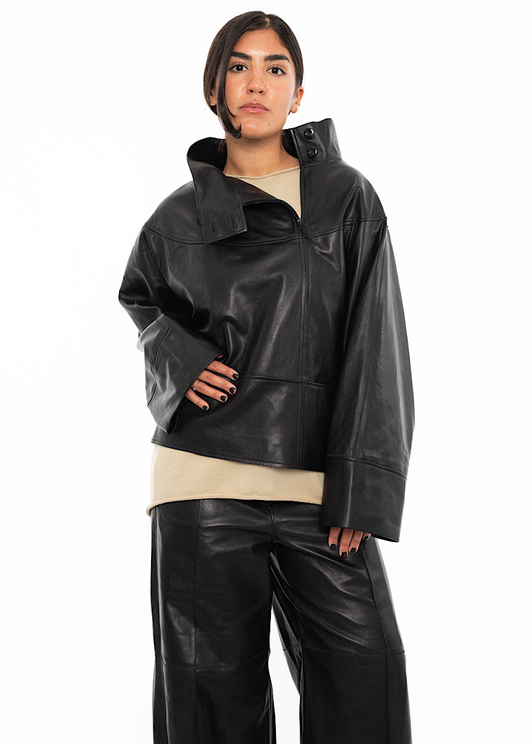 A model wearing an oversized Black Leather pullover with relaxed fit sleeves, black zipper detail and a distinctive open collar. Styled with matching wide leg trousers.