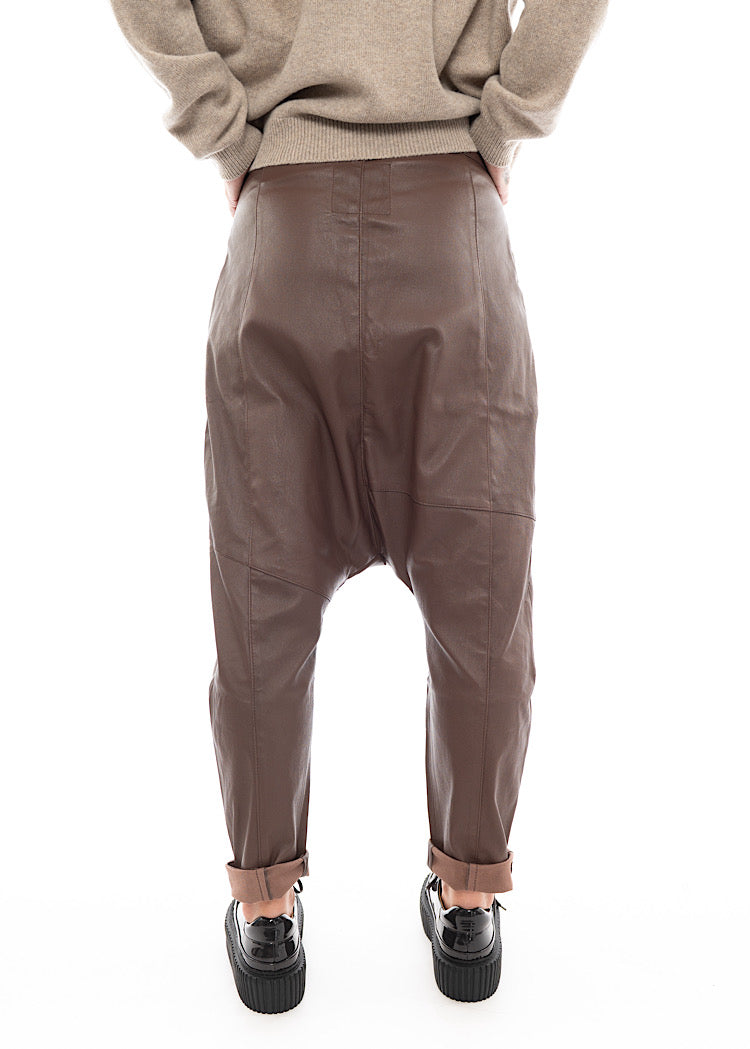 A Model standing facing the back wearing a Structured Drop-Crotch Brown Leather Trouser. Showing the seam-lines fro behind.