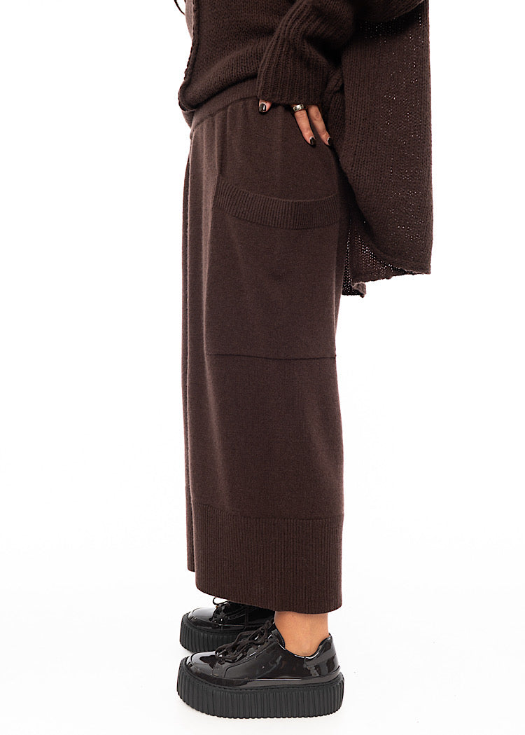 A Model standing to a side wearing a Brown Knitted Skirt with pockets. Showing the full length of the piece and the pockets.