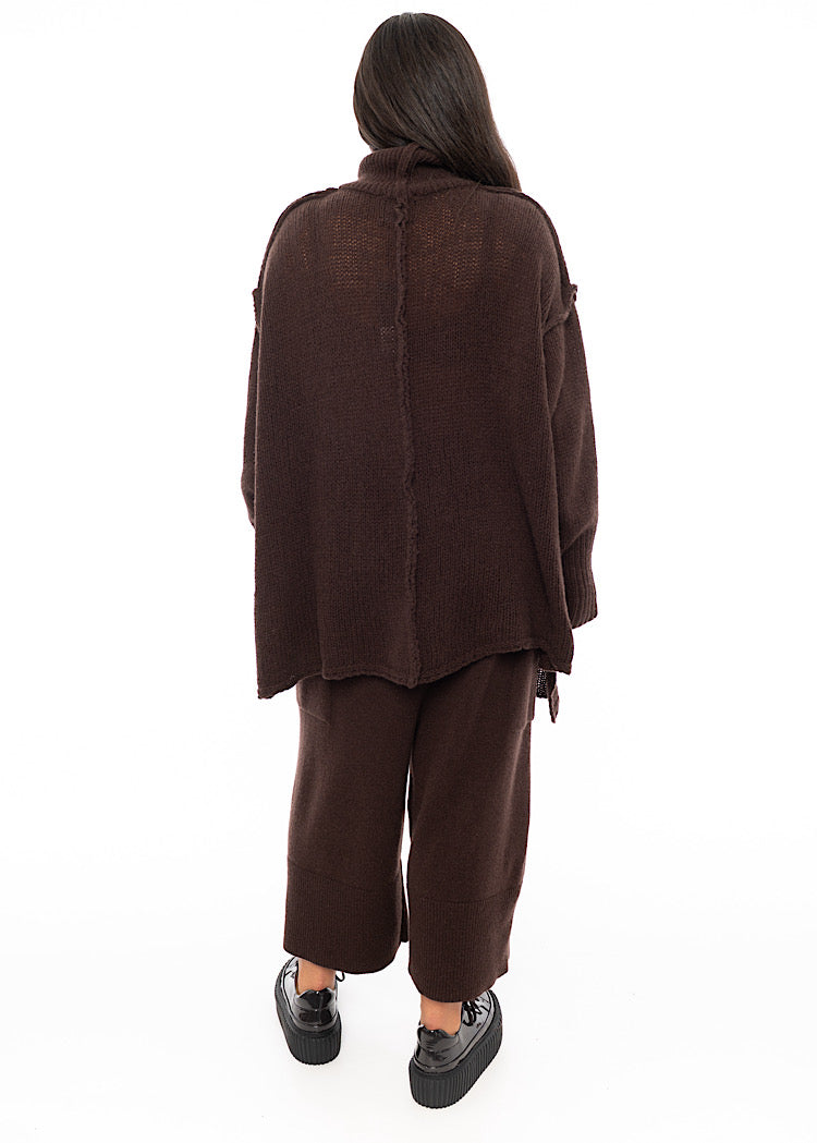 A Full frame photo of A Model standing facing the back wearing a Brown Oversized pullover. Showing the straight seam-lines and length.