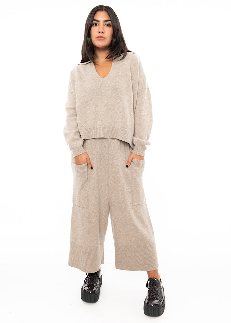 A full frame photo of A Model wearing a light beige knitted Trouser. Paired with a matching pullover and Brown patent Shoes.