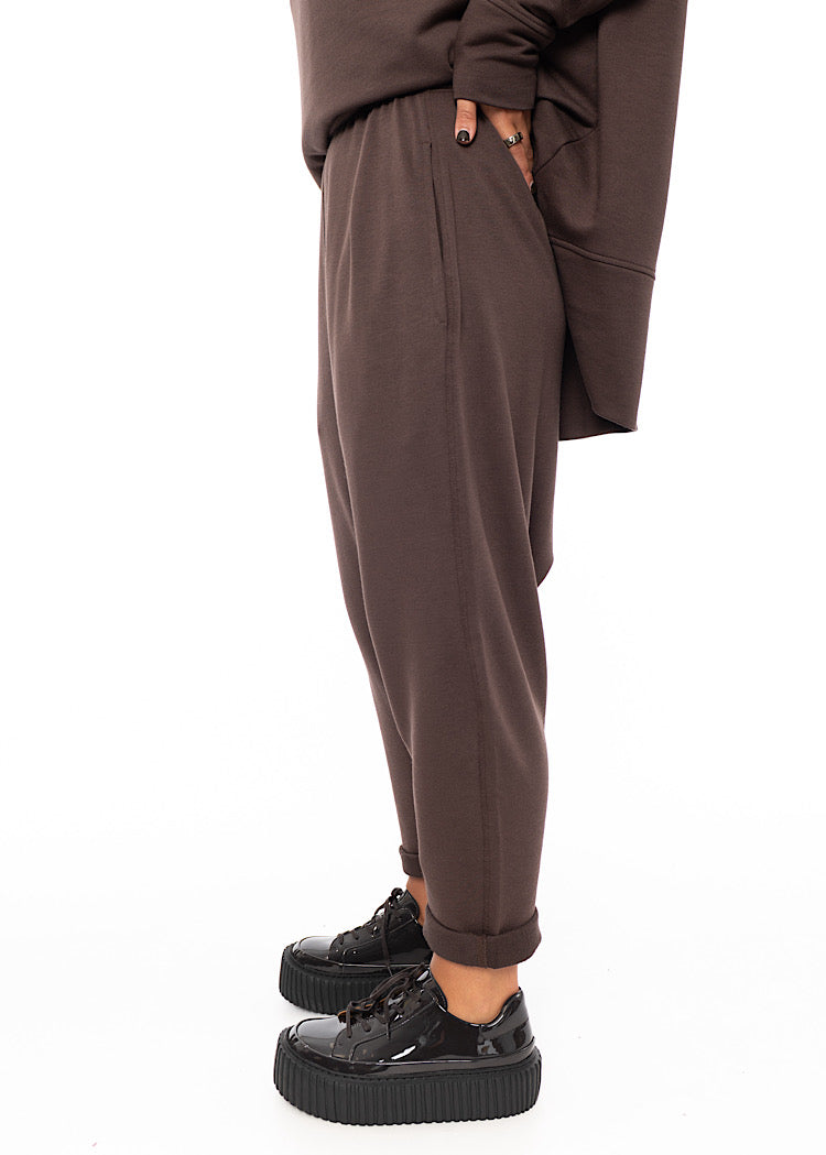 A Model standing to a side wearing a plum toned drop-crotch trouser with a slight rolled up effect. Showing the pockets and width of the trouser.