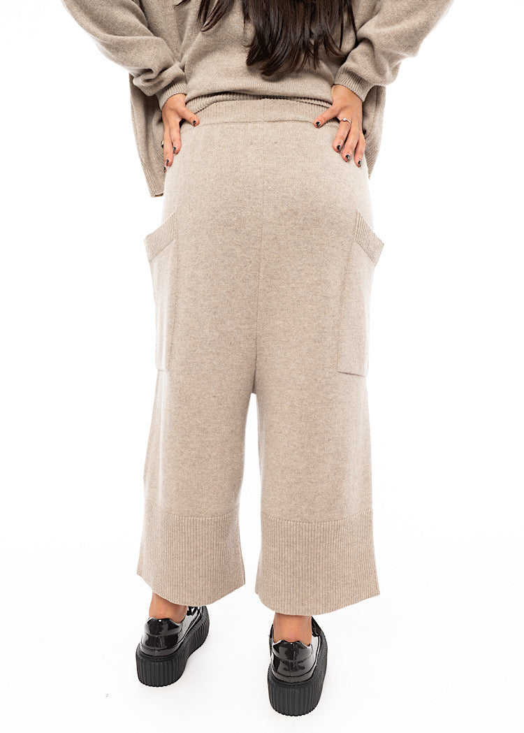 A Model wearing a light beige knitted Trouser. Paired with a matching pullover and Brown patent Shoes.Showing the fit from behind.