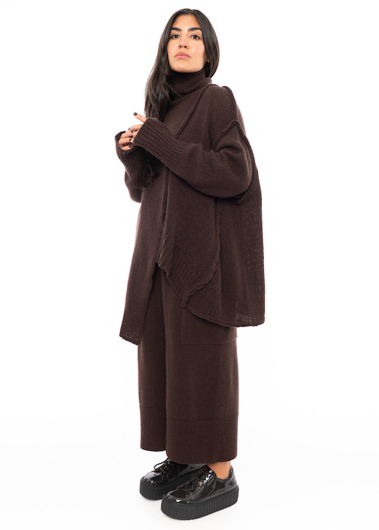 A Full frame photo of A Model standing to a side wearing a Brown Oversized pullover. Showing the pieces oversized length and Width.