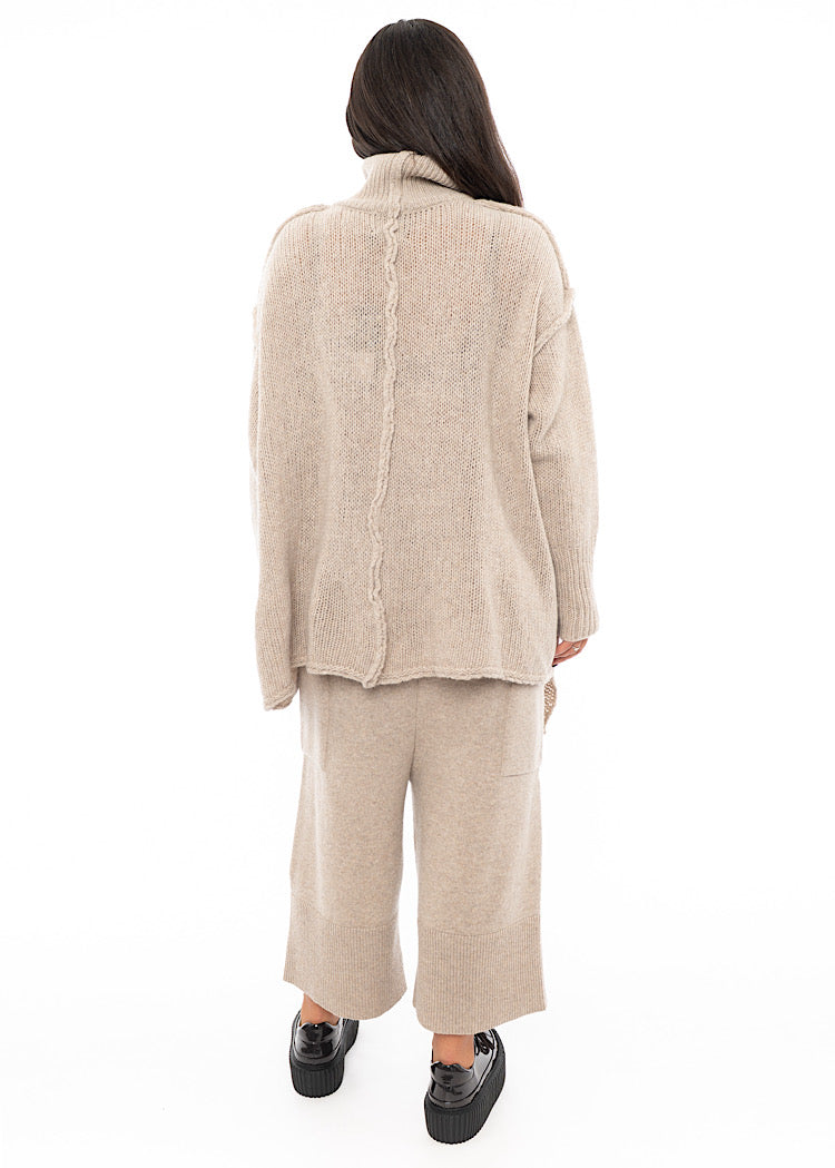 A full frame photo of A Model standing facing the back wearing a creme Knitted Pullover.Showing the Distinct seam-line throughout the piece.