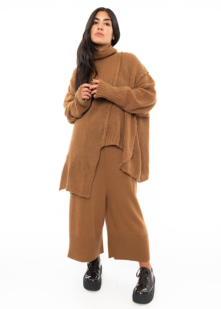 A Full Frame photo of A Model wearing an off-orange knitted Oversized Pullover. Paired with a matching coloured Trouser.