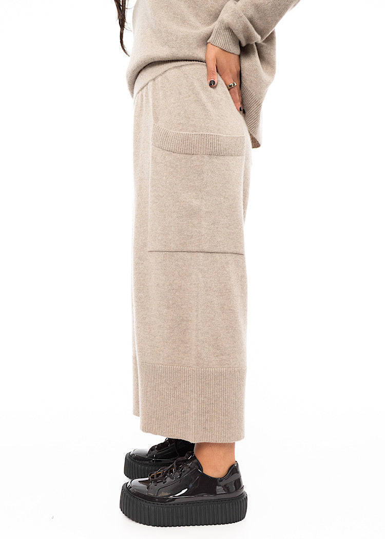 A Model wearing a light beige knitted Trouser. Paired with a matching pullover and Brown patent Shoes.Showing the full length of the piece.