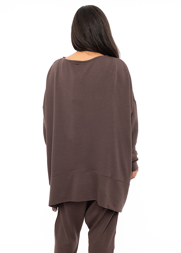 A Model standing facing the back wearing an oversized Plum toned Pullover, paired with a Matching Trouser. Showing the pieces long length.