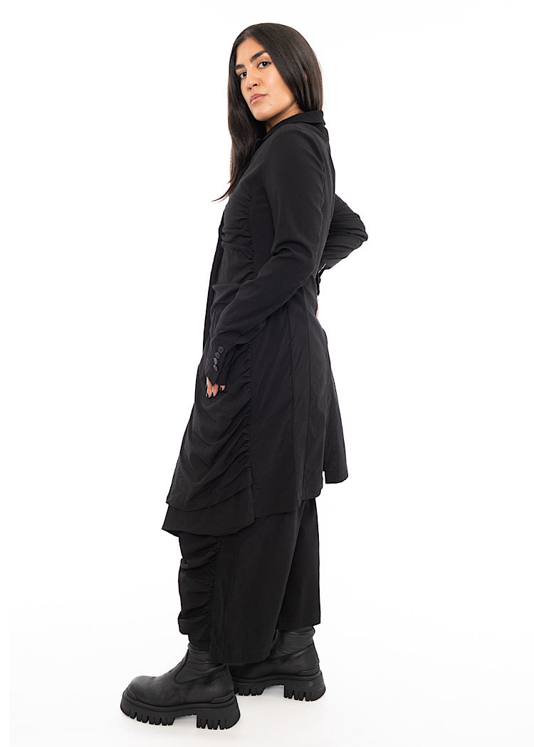 A Model standing to the side wearing a detailed techno coat and matching trousers, in a Black colour. Showing the fitted look from a different angle.
