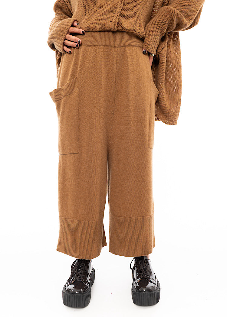 A Model wearing a Knitted pair of off-orange Trousers, paired with a matching pullover and patent shoes.Showing the front pockets.