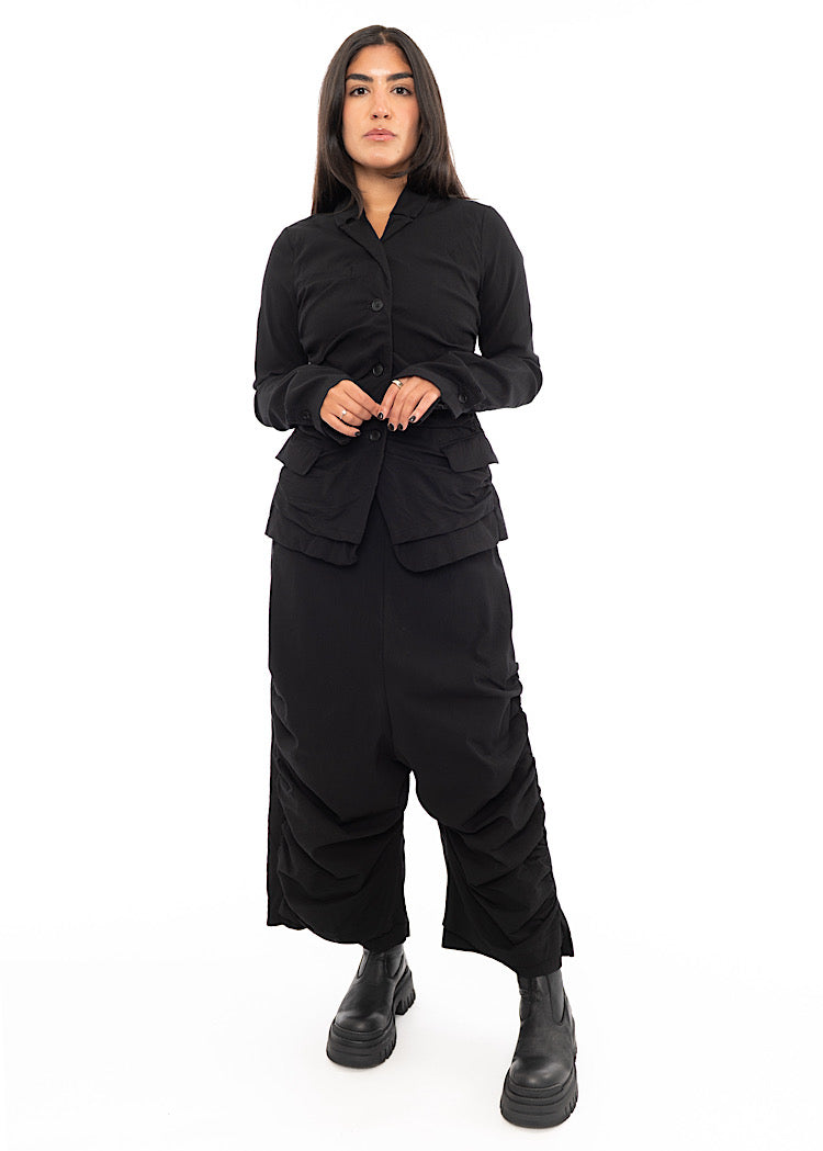 A full frame photo of A Model wearing a detailed techno Jacket and matching trousers, in a Black colour. Paired with a Black chunky boot.