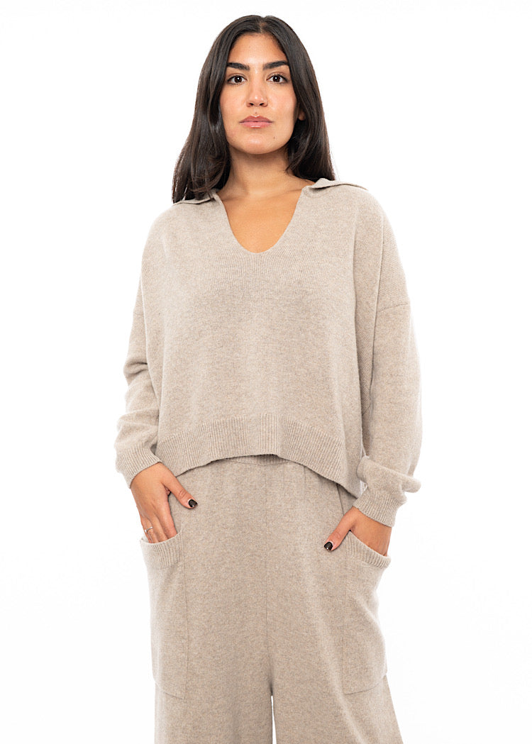 A Model wearing a light Beige knitted pullover with a ribbed hem and a deep V-neckline.Styled with a matching Trouser.