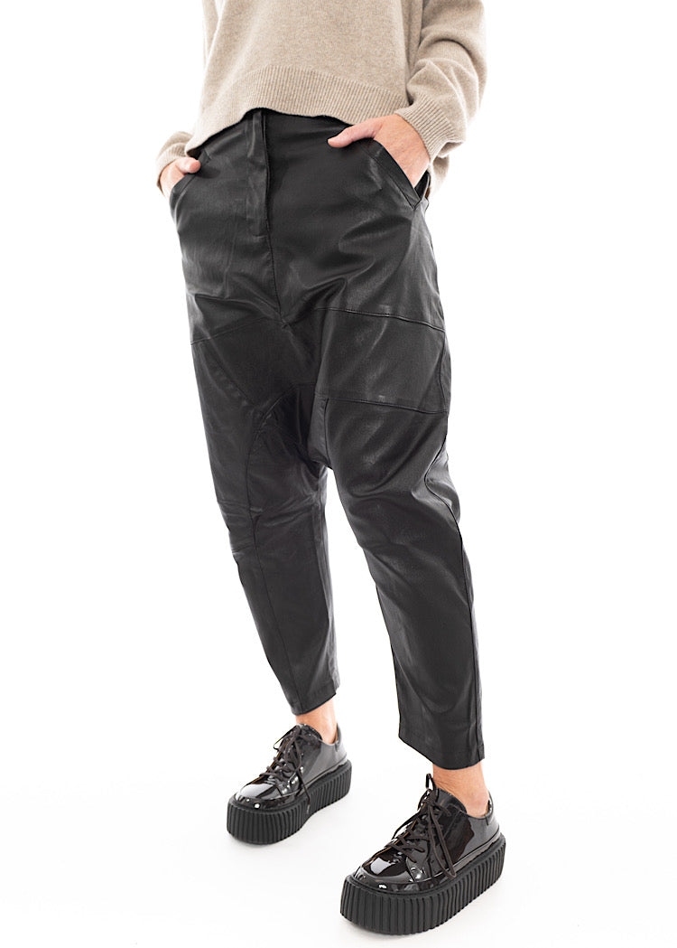 A Model wearing a Structured Drop-Crotch Black Leather Trouser, disclosing the pockets. Styled With a patent Black Shoe and a creme 