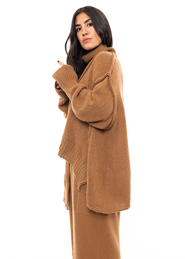 A Model standing to a side wearing a light brown knitted Oversized Pullover with distressed pieces dangling down. Showing the width and length of the piece.