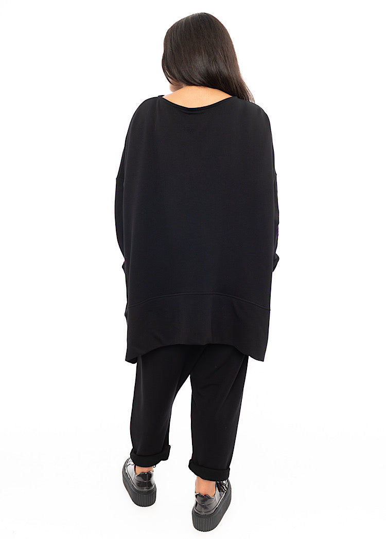 A Full frame photo of A Model standing to a side wearing a Black Oversized Pullover. Showing the behind dip neckline.